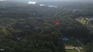 Aerial view of a wooded area with a few houses and a winding road in the foreground. A red star marks a specific spot among the trees. A large lake is visible in the background.