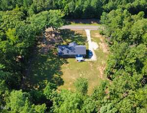 Aerial view of a single-story house with a dark roof, a driveway, and a small front porch, surrounded by trees and a grassy yard, with a road visible behind the property.