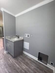 A laundry room with gray walls and flooring, a small gray cabinet with a sink and faucet, wall hookups for washer and dryer, and a metal dryer vent opening near the floor.