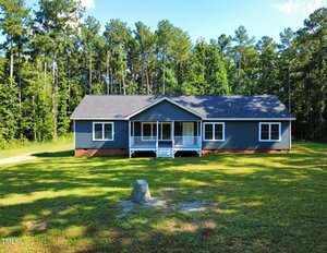 A single-story blue house with white trim sits on a grassy lawn at 1010 Pool Rock Road in Henderson, surrounded by tall trees. The house has a front porch with steps and railings, and multiple windows on the front facade.