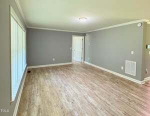 A bright, empty living room with light wood flooring, gray walls, white trim, and a ceiling light. Large windows line one wall, and a white door is visible at the far end of the room.