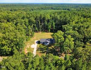 A single-story house with a gray roof is surrounded by dense green forest. There is a large clearing around the house, a driveway leading to the road, and a small patch of bare ground near the building.