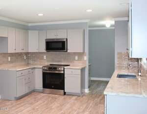 A modern kitchen with light gray cabinets, beige countertops, a stainless steel oven and microwave, beige tile backsplash, and wood-style flooring. The kitchen is empty and well-lit with ceiling lights.