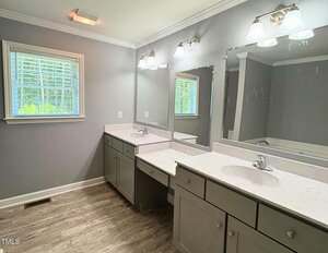 A bathroom with two sinks, gray cabinets, marble countertops, two mirrors, three light fixtures above the sinks, a window with blinds, and light-colored wood flooring.