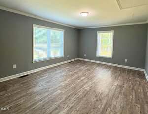 An empty room with gray walls, white trim, two large windows with blinds, wood-look flooring, and a ceiling light fixture. The room is well-lit with natural light.