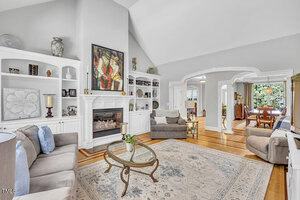 Living room with high vaulted ceiling, built-in white shelves, fireplace, neutral-toned furniture, a glass coffee table, light area rug, hardwood floors, and an archway leading to a dining room with large windows.