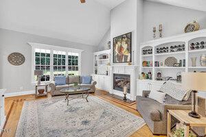 A bright living room with a high ceiling, a large rug, a glass coffee table, a sofa, an armchair, built-in white shelves with decor, a fireplace, and tall windows overlooking a green lawn.