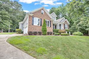 A brick and siding house with white trim, black shutters, a front porch, and attached garage, set on a sloped lawn with shrubs and trees surrounding the property.