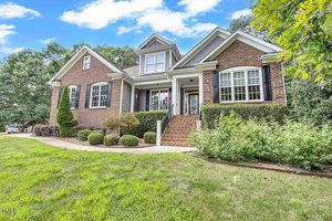 A two-story brick house with white trim, black shutters, and a front porch accessed by steps sits at 1001 Chatham Court in Youngsville. The grassy yard features neatly trimmed bushes and trees beneath a partly cloudy sky.
