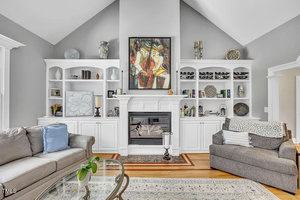 A living room with a high ceiling features a white fireplace and built-in shelves filled with books, art, and decor. Two sofas face each other, and a glass coffee table sits on a patterned rug in the center.
