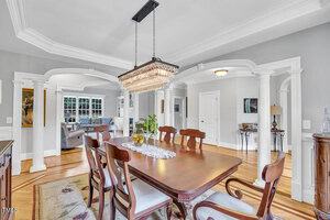 A formal dining room features a wooden table with six chairs, a crystal chandelier above, decorative white columns, hardwood floors, a rug, and archways leading to adjacent rooms with gray walls and crown molding.