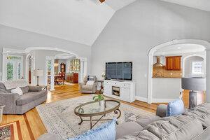 A spacious living room with light gray walls, hardwood floors, and vaulted ceilings. Furnishings include two gray sofas, a glass coffee table, a white TV console, and an area rug. Open archways lead to adjacent rooms.