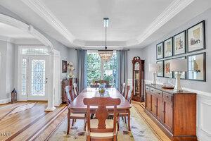 A formal dining room with a long wooden table, eight chairs, a chandelier, and a sideboard with lamps. There is a grandfather clock, framed artwork on the wall, large windows, and a glass-paneled front door.