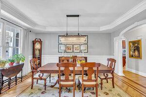 A formal dining room with a wooden table and six chairs, a patterned rug, a chandelier, a grandfather clock, framed artwork on a gray wall, and an arched doorway leading to another room.