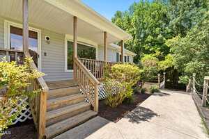 A wooden porch with steps and railing leads to the entrance of a light gray house. Shrubs and greenery line the walkway and the house is surrounded by trees under a clear sky.
