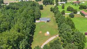Aerial view of a house surrounded by trees, with a large grassy yard, a small shed near the center, and a gravel driveway leading to the house. Several neighboring houses are visible in the distance.