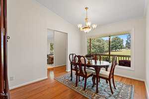 A dining room with a wooden table and six chairs on a decorative rug, a chandelier overhead, hardwood floors, neutral walls, and a large window with a view of grass and trees outside.