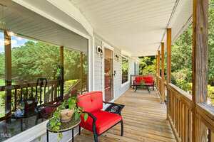 A wooden front porch with a white ceiling, two red cushioned chairs, a small round table with a potted plant, and wooden railings. Large windows reflect trees and greenery from the yard.