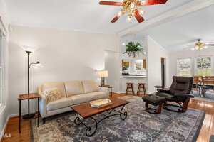 A living room with a beige sofa, wooden coffee table, and dark recliner on a patterned rug. Ceiling fans with lights are overhead, and the space opens to a kitchen and dining area with bar stools and large windows.