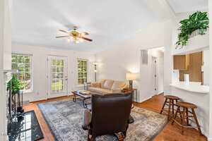 A bright living room with a ceiling fan, beige sofa, armchair, coffee table, and area rug. Large windows and double doors let in natural light. A breakfast bar with two stools is visible near the kitchen.