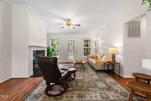 Living room with hardwood floors, a patterned rug, a brown recliner, a beige sofa, a coffee table, and a floor lamp. French doors and windows reveal greenery outside. Ceiling fan and fireplace are also visible.