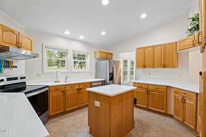 A spacious kitchen with wooden cabinets, white countertops, a stainless steel refrigerator, an electric stove, an island in the center, and multiple windows letting in natural light.