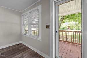 A small room with light-colored walls and wood-style flooring features two windows with white blinds and an open door leading to a covered wooden porch with a railing outside. Trees are visible in the background.