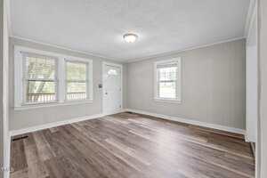 An empty room with light-colored walls, a white front door, three windows letting in natural light, a ceiling light fixture, and wood-style flooring in various shades of brown.
