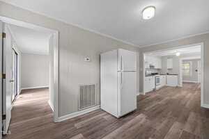 A kitchen with white cabinets and appliances, wood-look flooring, and beige paneled walls. An adjacent dining area and another room are visible through open doorways. Ceiling lights are turned on.