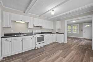 A kitchen with white cabinets, a white stove, and dark countertops. The floor is wood laminate, and the walls are light-colored. An open doorway leads to a living area with windows and a white front door.