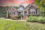A two-story brick house with white trim and black shutters at 1001 Chatham Court, Youngsville, featuring a front porch with steps, surrounded by shrubs and trees under a dramatic sunset sky.