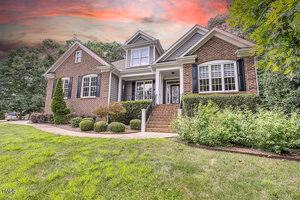 A two-story brick house with white trim and black shutters at 1001 Chatham Court, Youngsville, featuring a front porch with steps, surrounded by shrubs and trees under a dramatic sunset sky.