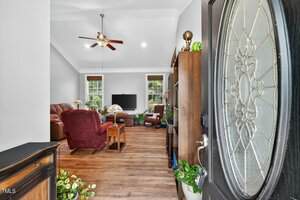 View of a living room from the open front door, showing wood flooring, armchairs, a sofa, a TV on a stand, houseplants, and a ceiling fan with lights. Large windows let in natural light.