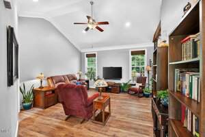 A living room with wood flooring, two red armchairs, a brown recliner sofa, wooden bookshelves, several potted plants, a flat-screen TV on the wall, and large windows letting in natural light.