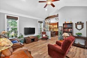 A living room with wood flooring, a brown recliner, a red armchair, a TV mounted on a wall, bookshelves, table lamps, and windows. The kitchen is visible through an open archway with decorative ironwork.