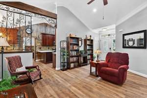 A living room with wood flooring, a red recliner, bookshelves, a side table with a lamp, an armchair, and decorative ironwork dividing the space from the kitchen. A ceiling fan hangs from the vaulted ceiling.