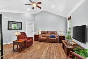 A living room with hardwood floors, gray walls, a brown reclining sofa, a matching recliner, a wall-mounted TV, two side tables with lamps, a large plant, a chess set, and a ceiling fan.