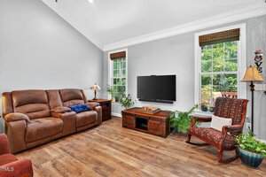 A living room with a brown reclining sofa, a patterned armchair, a wooden coffee table, a TV mounted on the wall, and large windows letting in natural light. Several green plants are placed around the room.
