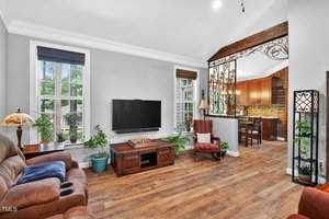 A living room with wood flooring, brown leather and red upholstered seating, a wall-mounted TV, plants, and a view into a kitchen with wood cabinets and decorative ironwork separating the spaces.
