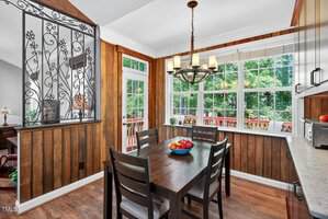 A dining area with wood-paneled walls, a dark wooden table and four chairs, a fruit bowl on the table, a large window, a glass door, and a decorative metal partition.