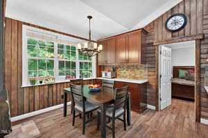 A kitchen with wood-paneled walls, a dining table with four chairs, a fruit bowl, large window, chandelier, wall clock, and cabinets. A doorway leads to a room with a bench and cushions.