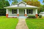 This charming single-story house at 807 N Main Street in Creedmoor features gray siding, white trim, a metal roof, and a welcoming covered front porch with brick columns. The home sits on a lush green lawn with trees and a shed in the background.