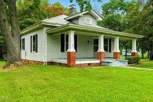 A single-story house with light gray siding, a metal roof, red brick foundation, a covered front porch with white columns, and a lawn with trees in the background.
