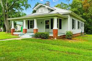 A single-story house with light gray siding, black shutters, a covered front porch supported by white columns with brick bases, and a neatly trimmed lawn surrounded by trees.