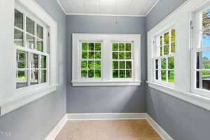 A small enclosed room with gray walls, three sets of white-trimmed windows, beige carpet flooring, and a ceiling light, providing a clear view of green trees outside.