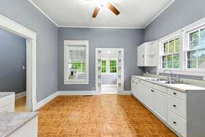 A spacious kitchen with gray walls, white cabinets, parquet wood flooring, a ceiling fan, and multiple windows letting in natural light. An adjacent room is visible through an open doorway.