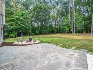 A backyard with a stone patio in the foreground, a circular fire pit area with four chairs on gravel, and a grassy lawn surrounded by tall trees in the background.