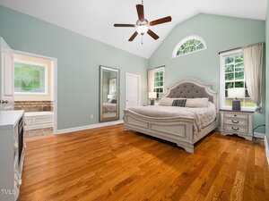 A spacious bedroom with light green walls, wood floors, a large bed with a tufted headboard, two windows with curtains, a ceiling fan, a wall mirror, and an open door leading to a bathroom.