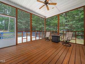 A screened-in porch with wooden floors and walls, two chairs, and a small table. A ceiling fan is overhead, and the porch overlooks a deck and a yard with trees.