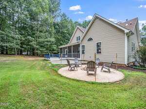 A beige house with a screened porch and above-ground pool in the backyard. A circular sand fire pit area with Adirondack chairs is set on the grass, bordered by trees in the background.